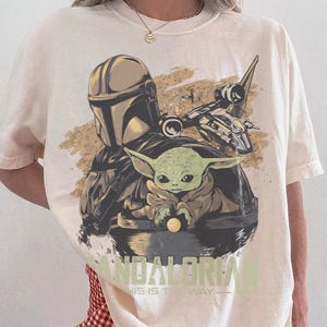 May include: Ivory-colored t-shirt featuring a graphic of The Mandalorian, Grogu, and a spaceship. The text "MANDALORIAN" and "THIS IS THE WAY" are printed below the image. The shirt is a Comfort Colors brand.
