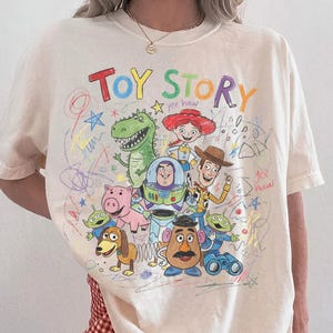Puede incluir: Camiseta de color marfil con un gr&aacute;fico colorido de los personajes de Toy Story, incluyendo a Woody, Buzz Lightyear y Rex. Las palabras "Toy Story" est&aacute;n escritas en una fuente juguetona encima de los personajes. La camiseta es de la marca Comfort Colors.