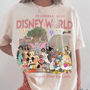 May include: Ivory-colored t-shirt featuring a graphic of Disney World in Florida. The design includes the words "Disney World" and "Magical Place On Earth" with various Disney characters and park icons. The shirt is a Comfort Colors brand.