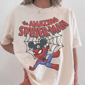 May include: Ivory-colored t-shirt featuring a cartoon Spider-Man character wearing a Mickey Mouse hat, hanging from a web. The words "The Amazing Spider-Man" are printed above the character in red and blue. The shirt is labeled "Comfort Colors Ivory".