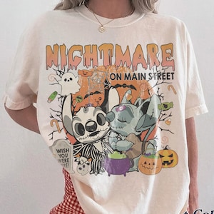Puede incluir: Camiseta de color marfil con un dise&ntilde;o de Halloween. El dise&ntilde;o presenta el texto "Nightmare on Main Street" y personajes de dibujos animados. La camiseta tambi&eacute;n tiene las palabras "Comfort Colors" e "Ivory" impresas.