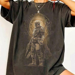 May include: A charcoal gray short-sleeve t-shirt featuring a graphic print of a helmeted warrior in armor holding a blaster. The design includes a circular emblem with intricate text surrounding the figure, rendered in muted gold and brown tones.