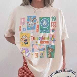 K&ouml;nnte beinhalten: Elfenbeinfarbenes T-Shirt mit einer bunten Disneyland-Grafik mit verschiedenen Zeichentrickfiguren und dem Text "Disneyland 1955". Das Shirt ist von Comfort Colors.
