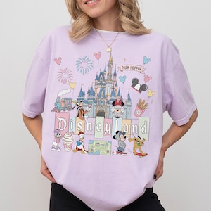 May include: A light purple t-shirt featuring a Disneyland graphic. The graphic includes the Disneyland castle, various Disney characters, fireworks, and the words "Disneyland" and "Park Hopper".