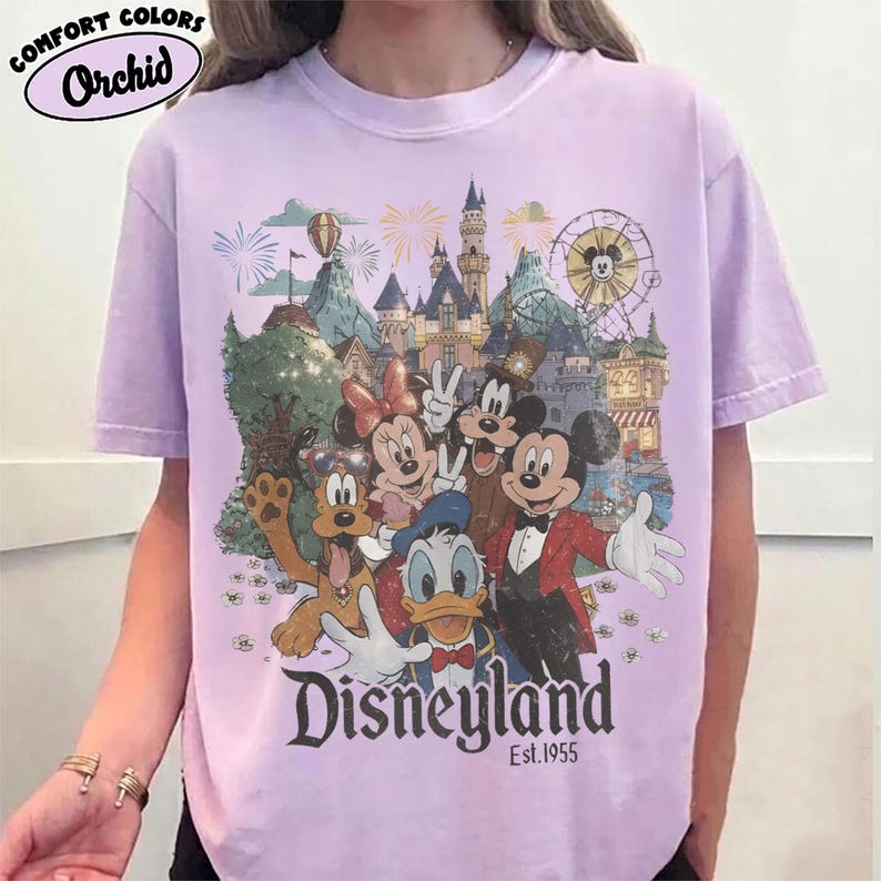May include: A light purple short-sleeve t-shirt featuring a graphic print of Mickey Mouse, Minnie Mouse, Donald Duck, Goofy, and Pluto in front of a Disneyland castle. The text reads Disneyland Est. 1955. The shirt is labeled Comfort Colors Orchid.