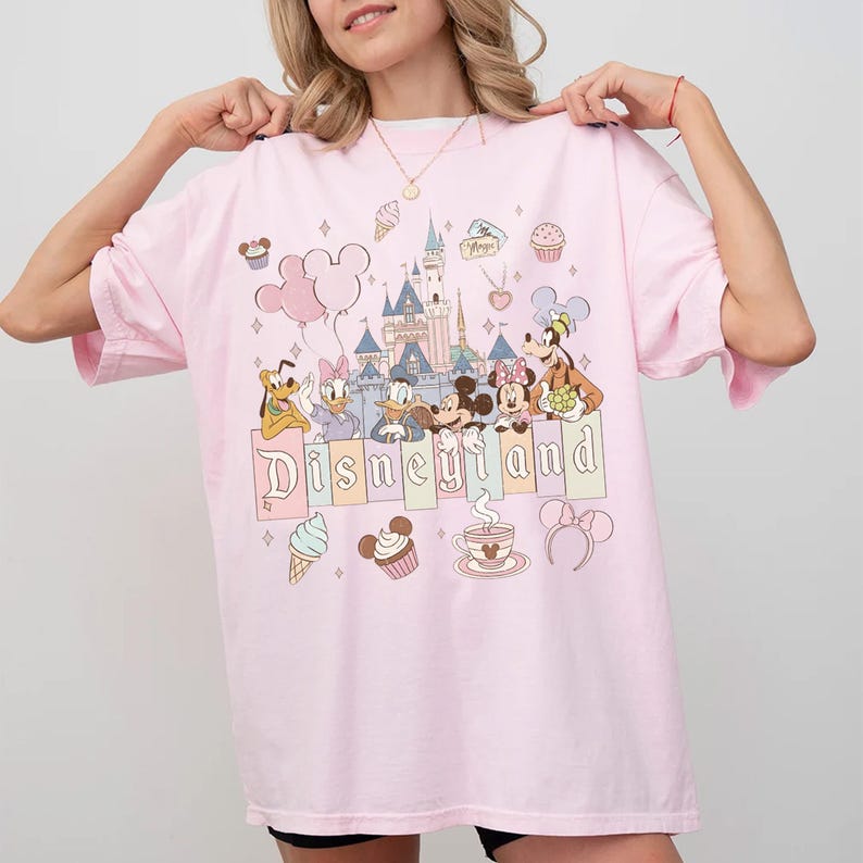May include: A pale pink t-shirt with a Disneyland design. The graphic includes cartoon characters, a castle, balloons, and the word "Disneyland". The shirt has short sleeves and a loose fit.
