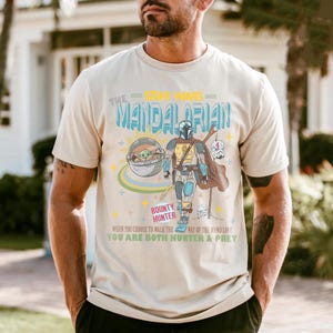 May include: Beige t-shirt featuring a graphic of The Mandalorian from Star Wars, with the text "The Mandalorian" and "Bounty Hunter". The design includes an illustration of the Mandalorian, Grogu, and other elements.