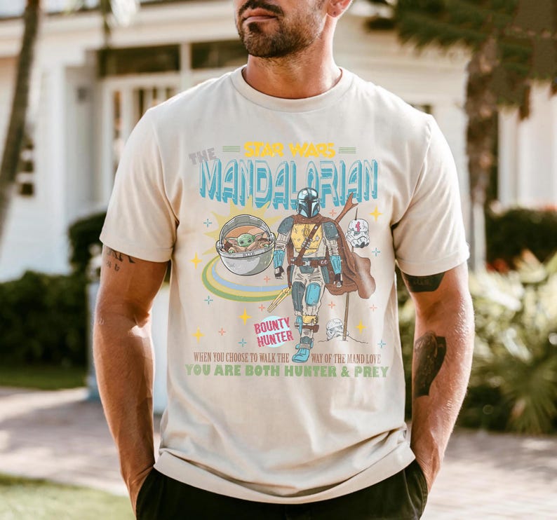 May include: Beige t-shirt featuring a graphic of The Mandalorian from Star Wars, with the text "The Mandalorian" and "Bounty Hunter". The design includes an illustration of the Mandalorian, Grogu, and other elements.