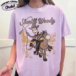 May include: A light purple short-sleeve t-shirt featuring a graphic of Sheriff Woody riding Bullseye the horse. The text "Sheriff Woody" is printed above the characters in a brown script font. The background graphic shows a desert landscape.