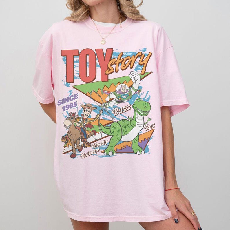 May include: A pale pink t-shirt with a vibrant Toy Story graphic. The design includes the words "TOY STORY" and "SINCE 1995", alongside illustrations of Woody, Buzz Lightyear, Rex, and Bullseye, characters from the film.