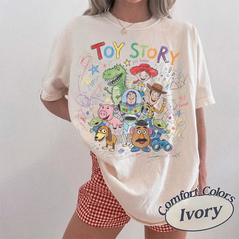 Puede incluir: Camiseta de color marfil con un gr&aacute;fico colorido de los personajes de Toy Story, incluyendo a Woody, Buzz Lightyear y Rex. Las palabras "Toy Story" est&aacute;n escritas en una fuente juguetona encima de los personajes. La camiseta es de la marca Comfort Colors.