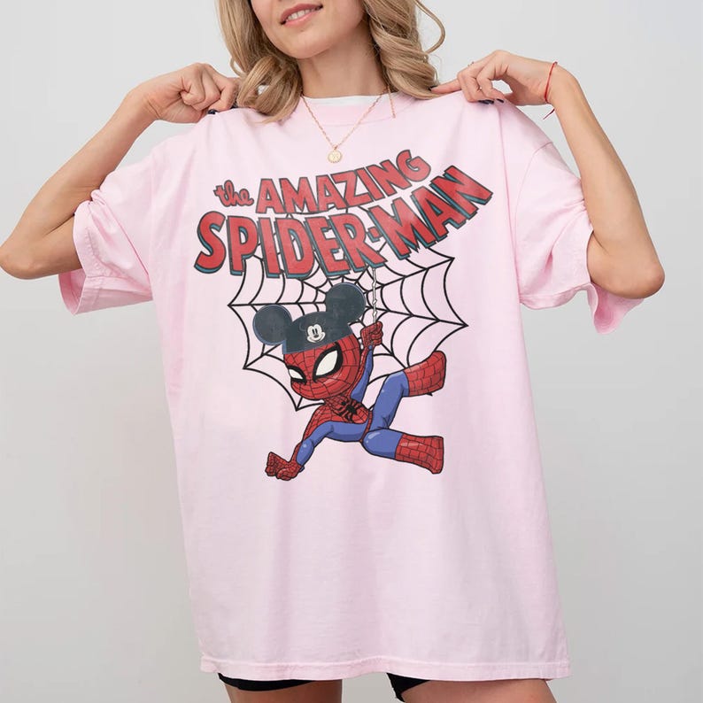 May include: A light pink t-shirt featuring a cartoon Spider-Man character wearing a Mickey Mouse hat, hanging from a web. The text "the AMAZING SPIDER-MAN" is printed across the top in red and white. The shirt has short sleeves.