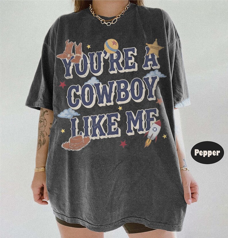 May include: A charcoal gray oversized t-shirt featuring the text YOU'RE A COWBOY LIKE ME in blue letters. The graphic includes illustrations of cowboy boots, a sheriff star, a ball, clouds, a rocket, and small stars in red, yellow, and white.