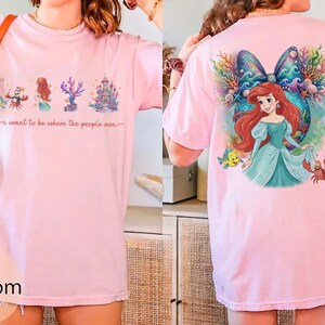 May include: Light pink t-shirt featuring a colorful graphic of a mermaid, castle, and coral. The front of the shirt has the text "I want to be where the people are". The back of the shirt has a large graphic of the mermaid with a bow.