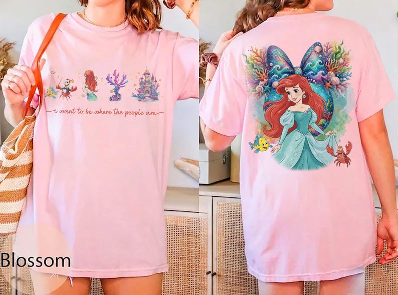 May include: Light pink t-shirt featuring a colorful graphic of a mermaid, castle, and coral. The front of the shirt has the text "I want to be where the people are". The back of the shirt has a large graphic of the mermaid with a bow.