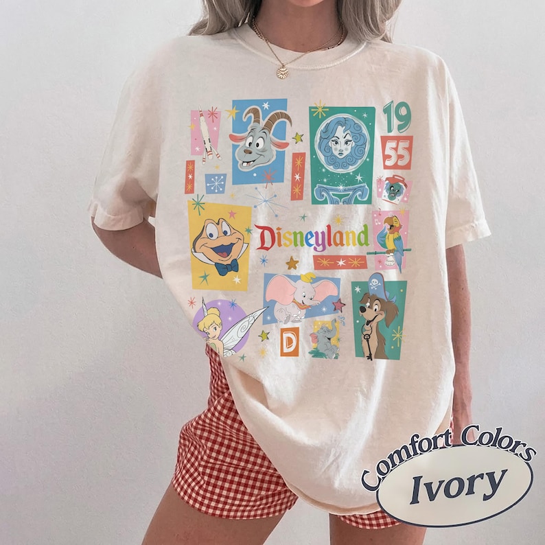 K&ouml;nnte beinhalten: Elfenbeinfarbenes T-Shirt mit einer bunten Disneyland-Grafik mit verschiedenen Zeichentrickfiguren und dem Text "Disneyland 1955". Das Shirt ist von Comfort Colors.