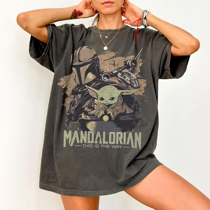 May include: Dark gray t-shirt featuring a graphic of the Mandalorian and Grogu from the Star Wars series. The design includes the text "MANDALORIAN" and "THIS IS THE WAY". The shirt has short sleeves and a relaxed fit.