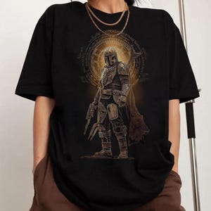May include: A black t-shirt featuring a graphic print of a armored character in a helmet and cape, holding a blaster. The design includes a circular golden emblem with intricate text surrounding the figure, rendered in shades of gold and brown.