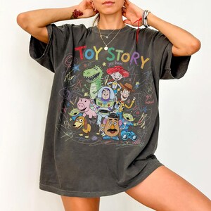 Puede incluir: Camiseta gris oscuro con las palabras "TOY STORY" en letras coloridas e ilustraciones de personajes de la pel&iacute;cula. El dise&ntilde;o incluye un dinosaurio, Buzz Lightyear, Woody y otros personajes, con el texto "yee haw".