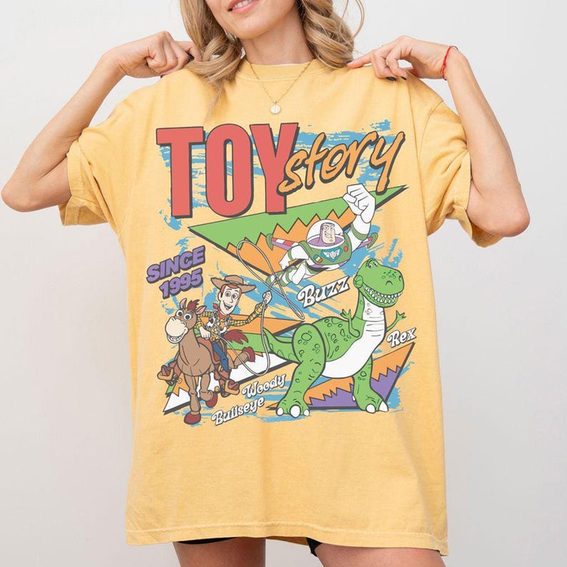 May include: Mustard yellow t-shirt with a Toy Story graphic. The design features Woody, Buzz Lightyear, and Rex, with the text "TOY Story" and "SINCE 1995". The shirt has a vintage, slightly faded appearance.