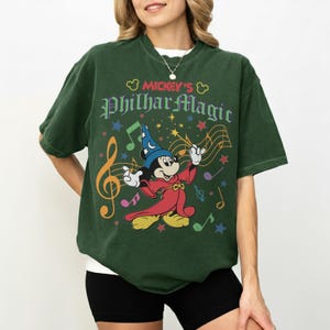 May include: Dark green t-shirt featuring Mickey Mouse as the Sorcerer's Apprentice from Fantasia. The graphic includes musical notes, stars, and the text "Mickey's PhilharMagic" in a colorful design. The shirt has a relaxed fit.