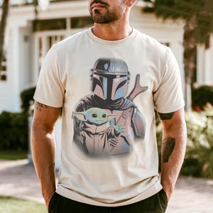 May include: A cream-colored short-sleeve t-shirt featuring a graphic print of a helmeted warrior in silver armor holding a small green creature with large ears and a tan robe. The design is centered on the chest of the garment.