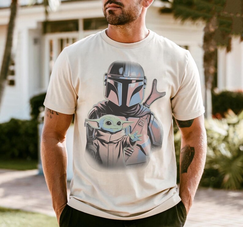 May include: A cream-colored short-sleeve t-shirt featuring a graphic print of a helmeted warrior in silver armor holding a small green creature with large ears and a tan robe. The design is centered on the chest of the garment.