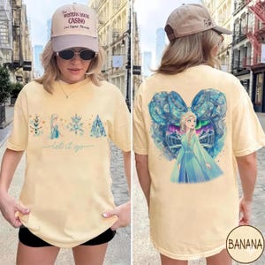 May include: A light yellow t-shirt featuring a front graphic of a snowman, a princess, snowflakes, and a castle with the text "let it go". The back of the shirt has a graphic of a princess with a bow and aurora borealis.