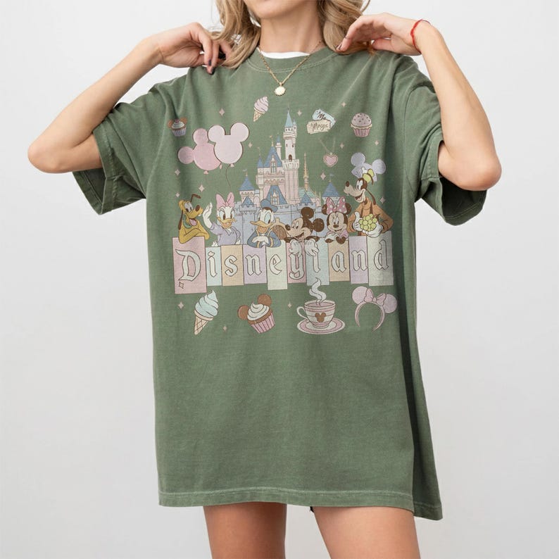 May include: Olive green t-shirt with the word "Disneyland" and a castle graphic, featuring cartoon characters. The shirt includes images of Mickey Mouse, Minnie Mouse, Donald Duck, and other Disney characters. The shirt has a relaxed fit.