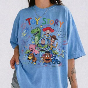 Puede incluir: Camiseta azul claro con un estampado colorido de los personajes de Toy Story, incluyendo a Woody y Buzz Lightyear. Las palabras "Toy Story" est&aacute;n escritas sobre los personajes. La camiseta tiene un corte holgado.