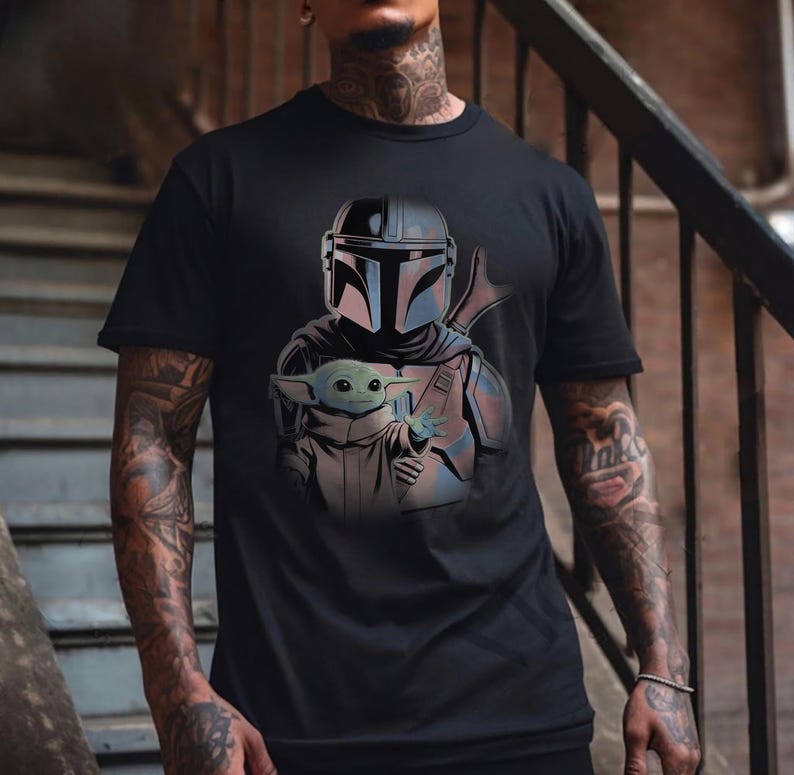 May include: A black short-sleeved t-shirt featuring a graphic print of a helmeted warrior in silver armor holding a small green creature with large ears and eyes. The design is centered on the chest, showcasing detailed shading and character features.