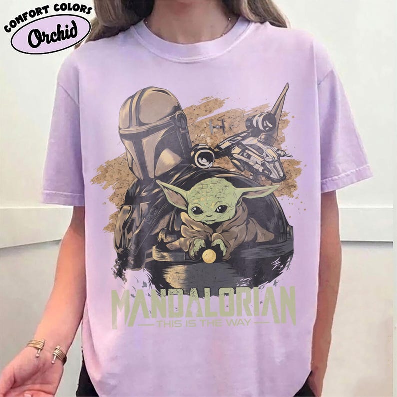 May include: Orchid Comfort Colors t-shirt in a light purple hue. The shirt features a graphic of The Mandalorian, Grogu, and a spaceship. The text "MANDALORIAN" and "THIS IS THE WAY" are printed below the characters.
