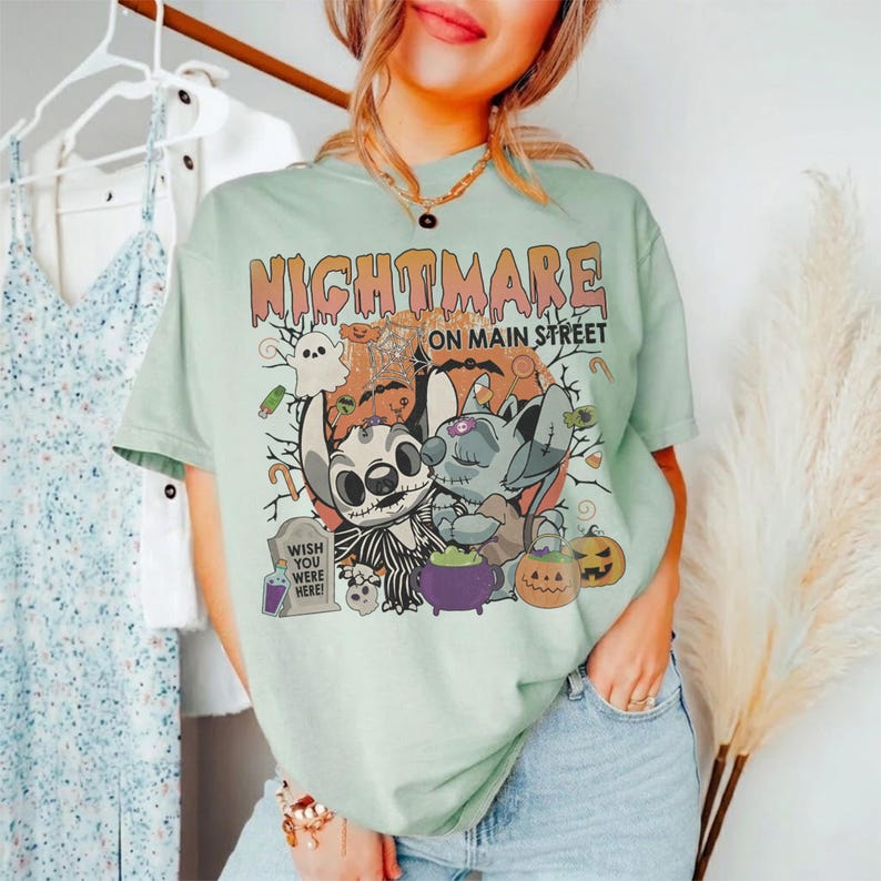 Puede incluir: Camiseta verde claro con un dise&ntilde;o de Halloween. El dise&ntilde;o incluye el texto "NIGHTMARE ON MAIN STREET", personajes de dibujos animados, calabazas y una l&aacute;pida con el texto "WISH YOU WERE HERE!"