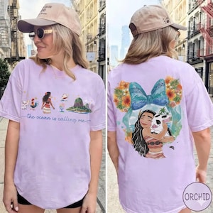May include: Light purple t-shirt featuring a colorful graphic of a cartoon character with a pig, surrounded by flowers and a blue bow. The front of the shirt has a landscape design with the text "the ocean is calling me".
