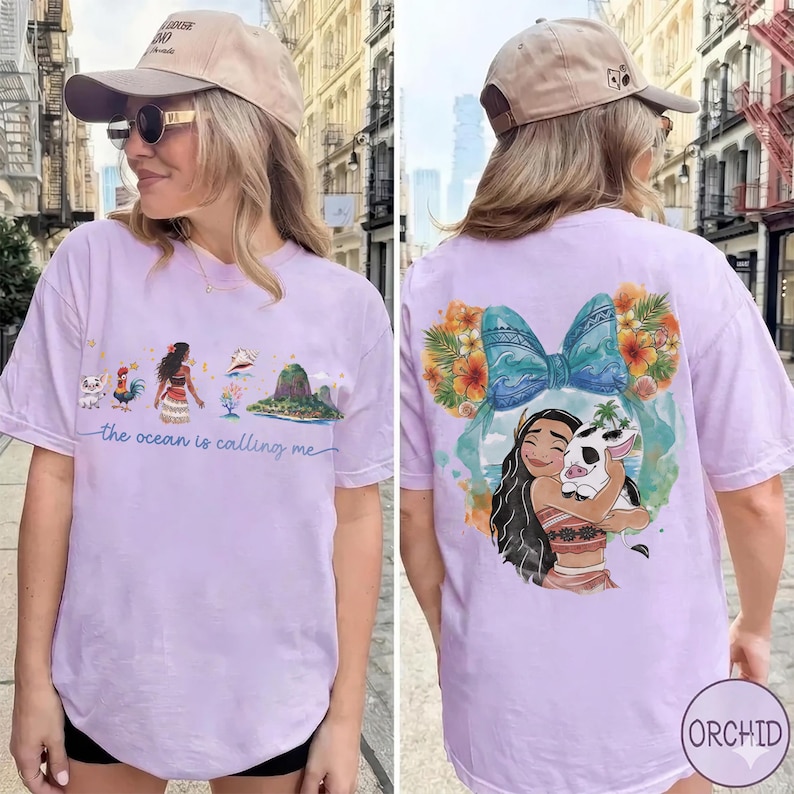 May include: Light purple t-shirt featuring a colorful graphic of a cartoon character with a pig, surrounded by flowers and a blue bow. The front of the shirt has a landscape design with the text "the ocean is calling me".
