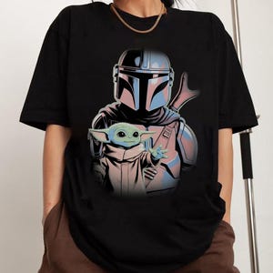 May include: A black short-sleeved t-shirt featuring a graphic print of a helmeted character in silver and blue armor standing behind a small green creature with large ears and eyes, wearing a tan robe and reaching out with one hand.