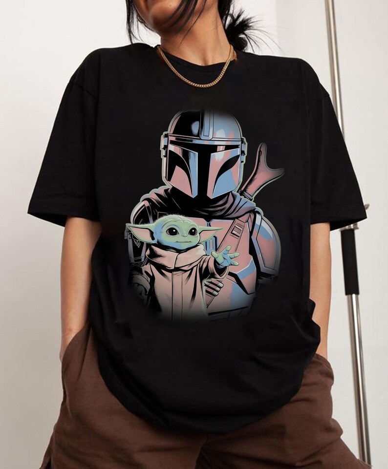 May include: A black short-sleeved t-shirt featuring a graphic print of a helmeted character in silver and blue armor standing behind a small green creature with large ears and eyes, wearing a tan robe and reaching out with one hand.