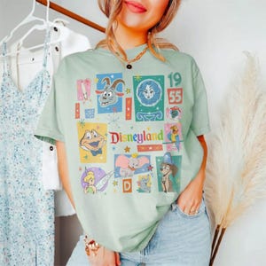 K&ouml;nnte beinhalten: Hellgr&uuml;nes T-Shirt mit einer farbenfrohen Grafik von Disneyland-Charakteren und dem Text "Disneyland 1955". Das Design umfasst Bilder einer Ziege, eines Frauengesichts, eines Frosches, Dumbo und anderer Zeichentrickfiguren.