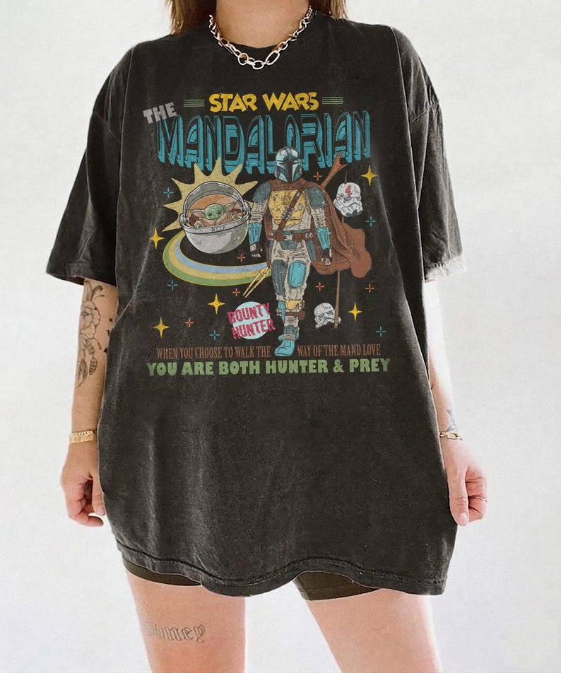 May include: Dark gray t-shirt featuring a graphic of The Mandalorian from Star Wars. The design includes the Mandalorian, Grogu, and text that reads "BOUNTY HUNTER" and "YOU ARE BOTH HUNTER & PREY".