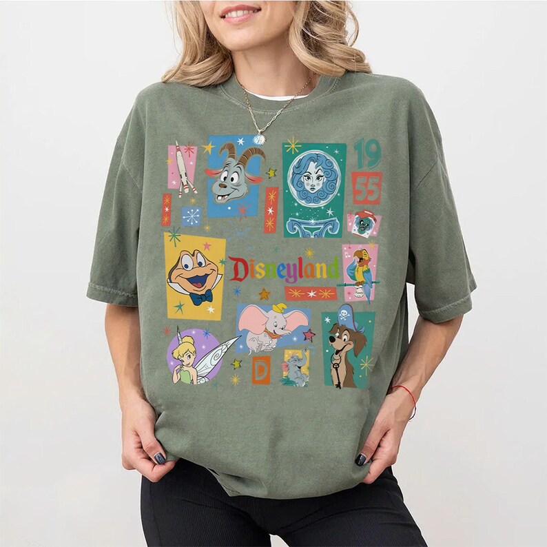 K&ouml;nnte beinhalten: Salbeigr&uuml;nes T-Shirt mit einer farbenfrohen Collage aus Vintage-Disneyland-Charakteren und Grafiken. Das Design umfasst Bilder von Dumbo, Tinkerbell und anderen klassischen Charakteren, mit dem Text "Disneyland" und dem Jahr "1955".