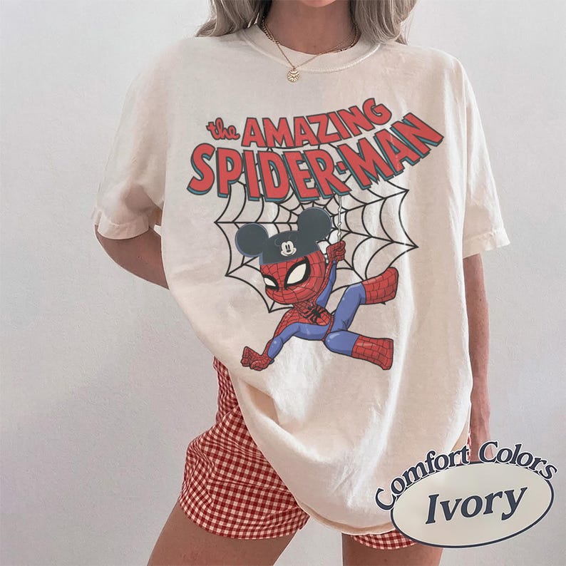 May include: Ivory-colored t-shirt featuring a cartoon Spider-Man character wearing a Mickey Mouse hat, hanging from a web. The words "The Amazing Spider-Man" are printed above the character in red and blue. The shirt is labeled "Comfort Colors Ivory".
