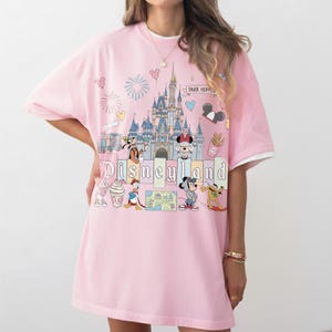 May include: Pink t-shirt with a Disneyland graphic featuring a castle, fireworks, and cartoon characters. The shirt has a white trim on the sleeves and collar. The text "Disneyland" is printed across the front.
