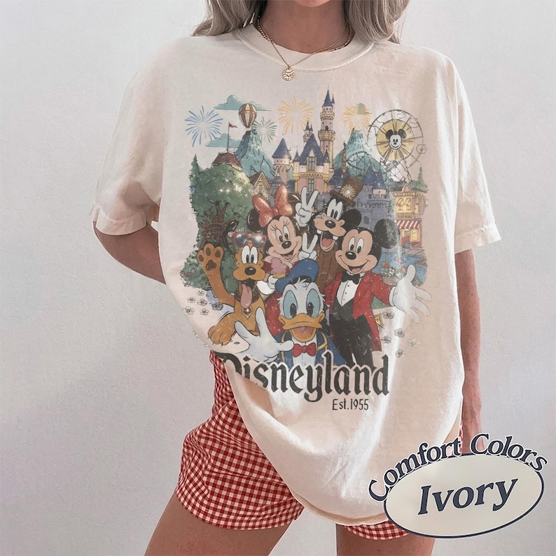 May include: An ivory short-sleeve t-shirt featuring a graphic print of Mickey Mouse, Minnie Mouse, Donald Duck, and Pluto in front of a castle and Ferris wheel. The text reads Disneyland Est. 1955. A small oval label in the corner says Comfort Colors Ivory.