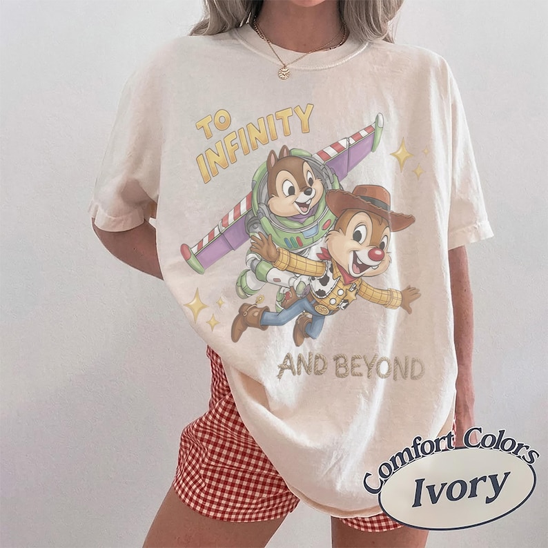 May include: An ivory short-sleeve t-shirt featuring a graphic of two chipmunks dressed as space and cowboy characters. The text reads TO INFINITY AND BEYOND. The shirt is paired with red and white checkered shorts. A label indicates Comfort Colors Ivory.