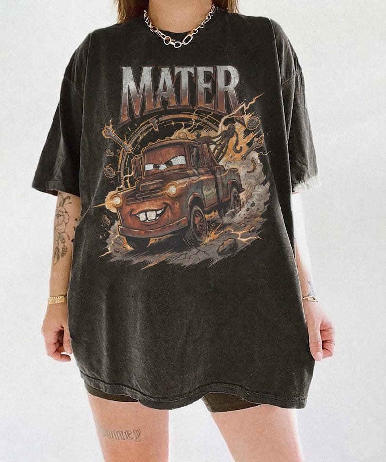 Może przedstawiać: Czarny t-shirt z grafiką Złomka z filmu Auta. Projekt zawiera słowo "MATER" w postarzonej czcionce nad postacią. Obraz przedstawia Złomka wzbijającego kurz i kamienie.