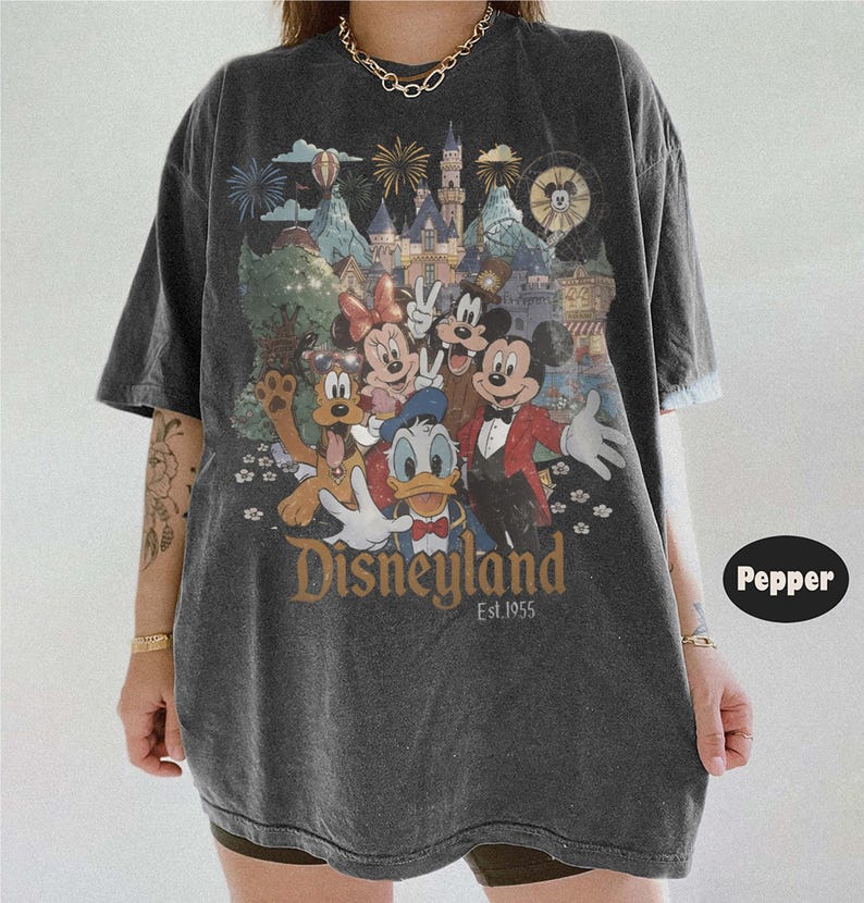 May include: A charcoal gray short-sleeve t-shirt featuring a graphic print of Mickey Mouse, Minnie Mouse, Donald Duck, Goofy, and Pluto in front of a castle. The text reads Disneyland Est. 1955. The shirt has a crew neckline and a relaxed, loose fit.
