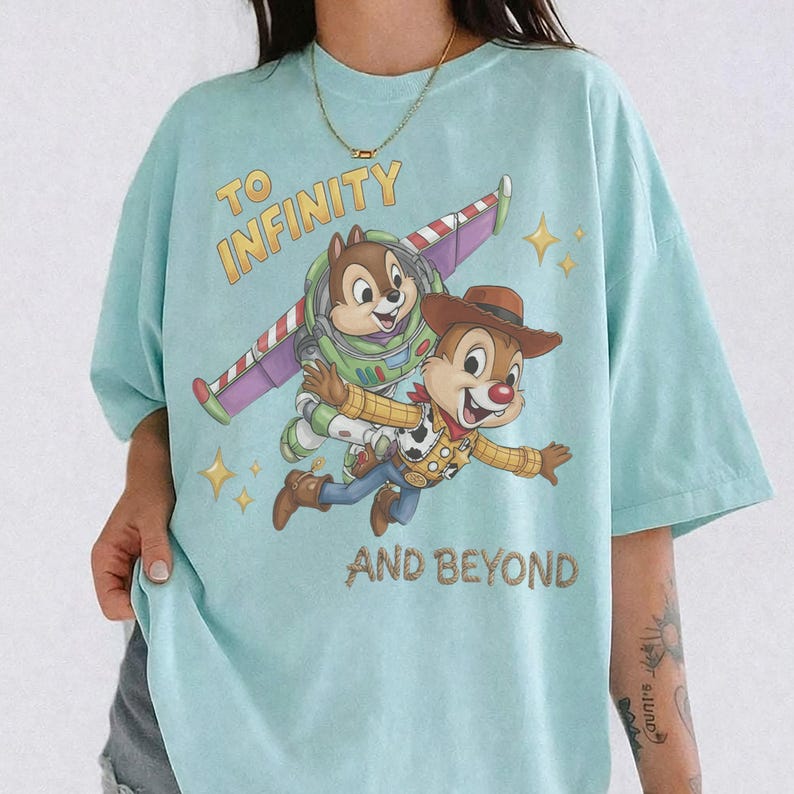 May include: A light blue t-shirt featuring a graphic of two chipmunks dressed as space and cowboy characters. One chipmunk wears a green and purple space suit with wings, while the other wears a cowboy hat and vest. The text reads TO INFINITY AND BEYOND.