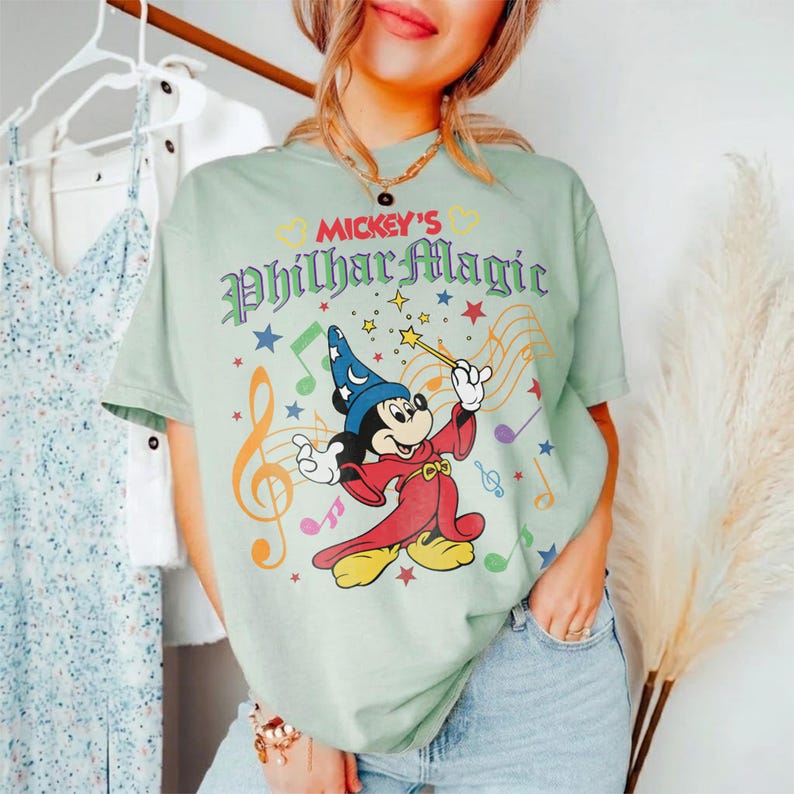 May include: A light green t-shirt featuring Mickey Mouse in a wizard costume with the text "Mickey's PhilharMagic". The graphic includes musical notes and stars. The shirt is a casual, comfortable style.