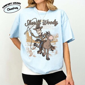 May include: A light blue short-sleeve t-shirt featuring a graphic print of Sheriff Woody riding a horse with a lasso. The text "Sheriff Woody" is printed above the character in a brown script font. A "Comfort Colors Chambray" logo is visible.
