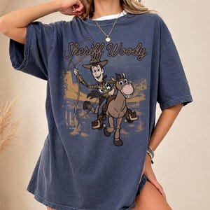 May include: A slate blue short-sleeve t-shirt featuring a graphic print of Sheriff Woody riding a horse. The design includes a lasso, a desert landscape background, and the text Sheriff Woody in a brown script font across the upper chest area.